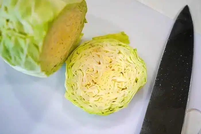 A halved cabbage is placed on a white surface next to a large kitchen knife, ready to be transformed into delicious baked cabbage steaks. The inner layers of the cabbage are visible, showcasing its tightly packed, light green and white leaves.