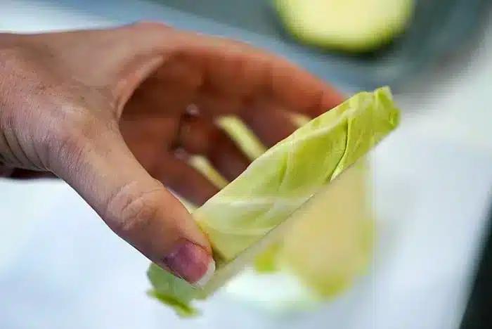 A close-up image of a hand holding a thin slice of green cabbage. The hand is grasping the slice lightly, showcasing its thickness and layers, reminiscent of preparing baked cabbage steaks. The background is blurred with another piece of cabbage visible.