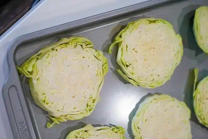Slices of cabbage arranged on a baking sheet, ready for roasting or baking into delicious baked cabbage steaks. The cabbage is cut into thick rounds, showing the layers of leaves within each slice. The baking sheet is placed on a countertop.