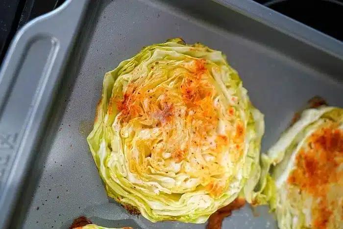 A close-up of a baked cabbage steak resting on a baking sheet. The seasoned cabbage is lightly browned, especially around the edges. The baking sheet has a slightly raised border.
