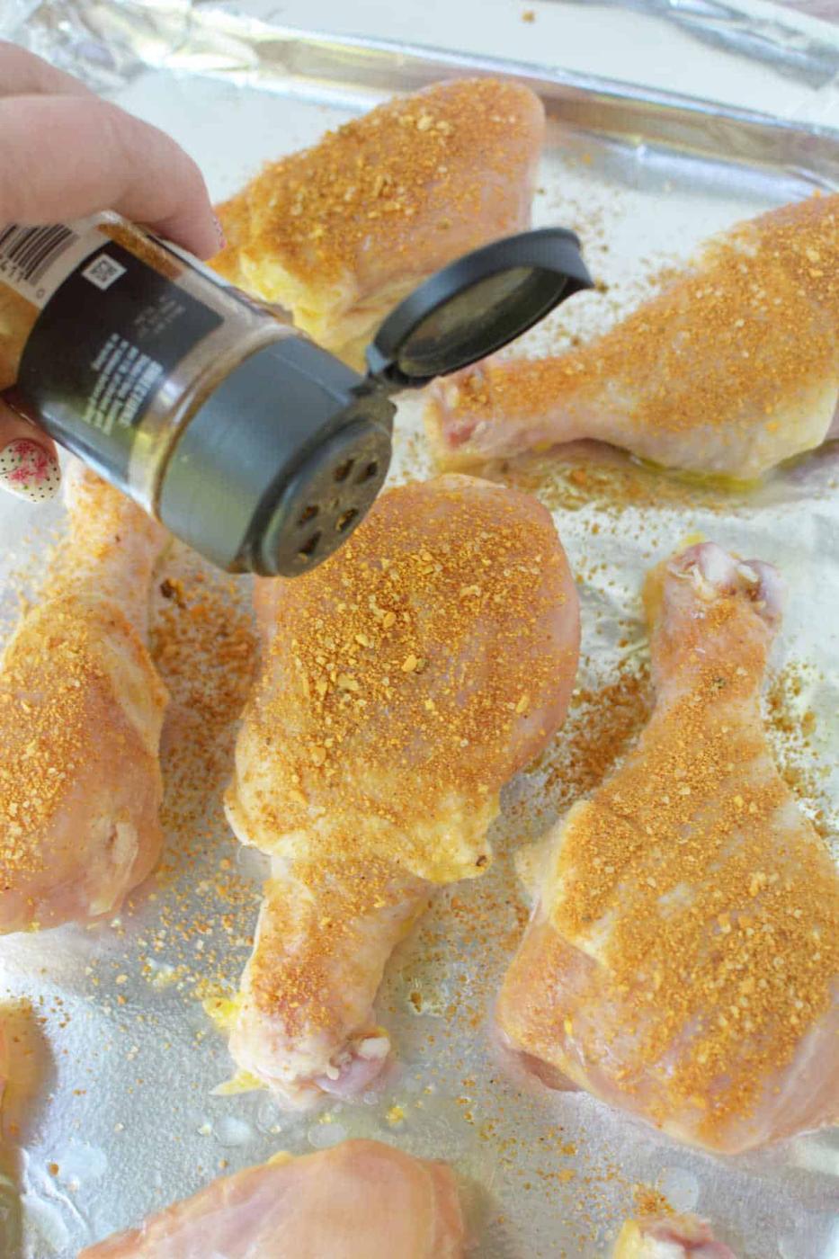 A hand sprinkles seasoning onto raw chicken drumsticks on a foil-lined baking sheet, preparing them to become flavorful baked chicken legs.