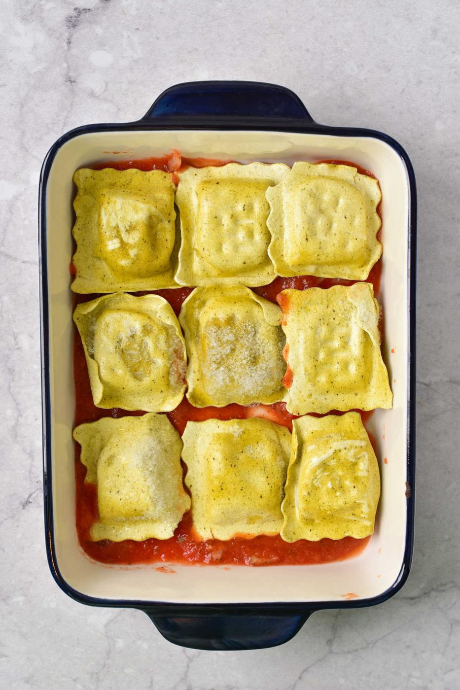 Nine pieces of uncooked baked frozen ravioli rest on a layer of tomato sauce in a rectangular baking dish.