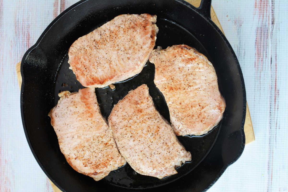 Four seasoned pork chops with apples cooking in a black cast iron skillet on a light wooden surface.