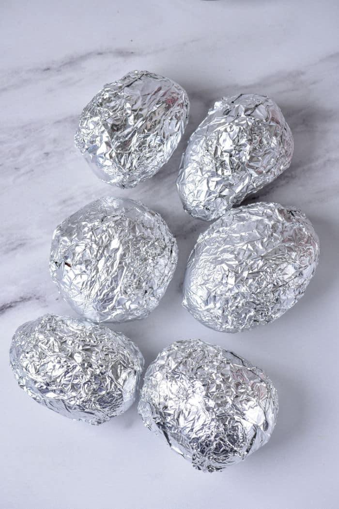 Six baked red potatoes are wrapped in crumpled aluminum foil and placed on a white marble surface.