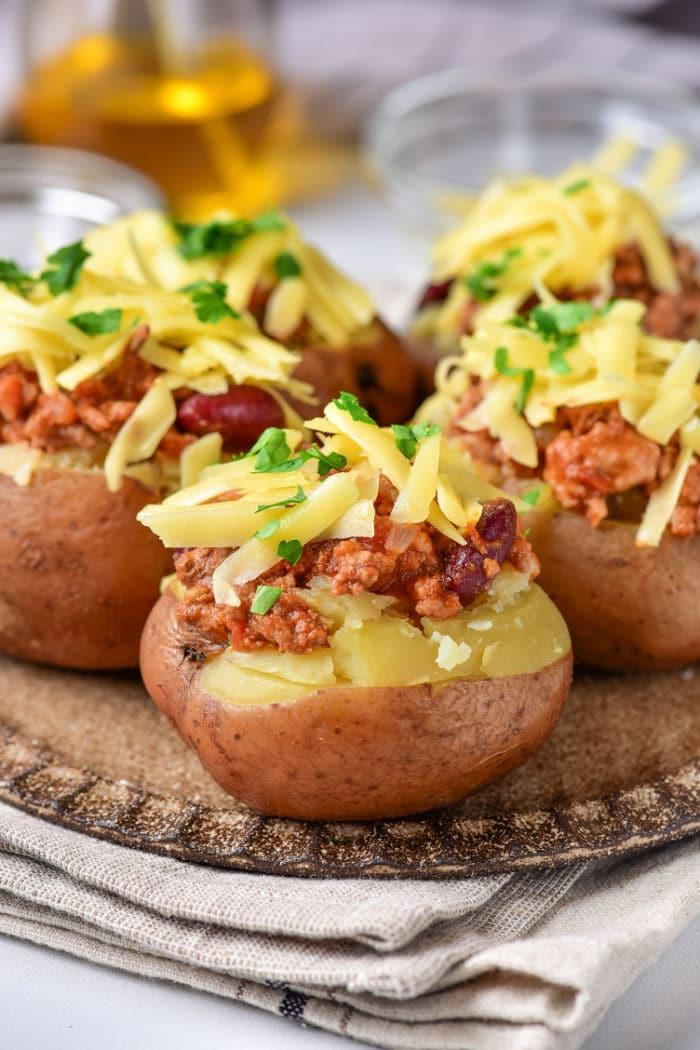 Three baked red potatoes topped with chili, shredded cheese, and chopped parsley are displayed on a rustic, round wooden plate. The plate is placed on a folded beige cloth. In the background are glass containers, slightly out of focus.