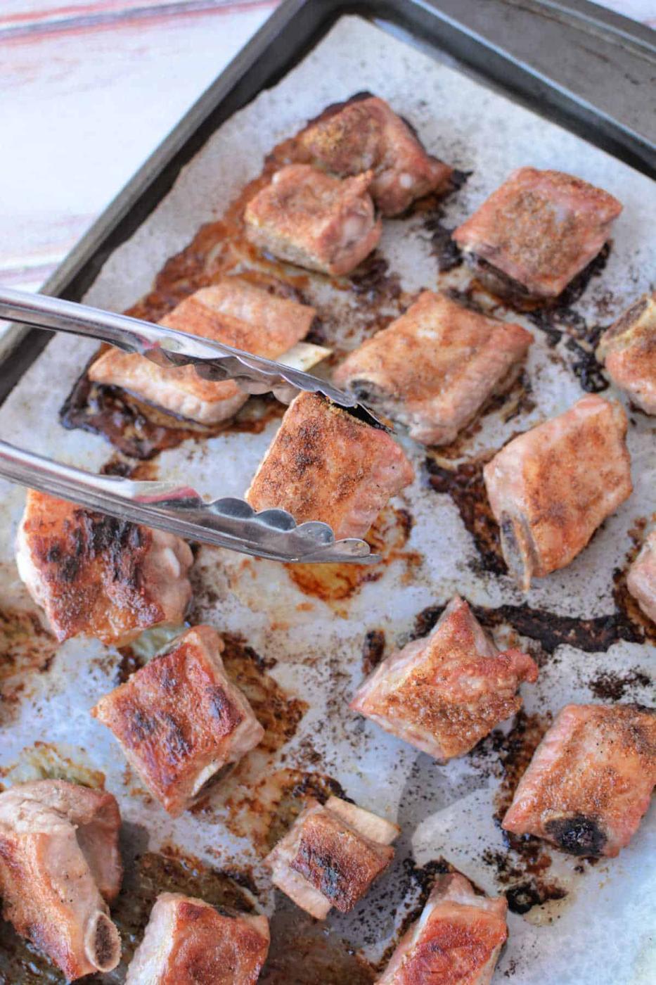 Tongs holding a piece of seasoned, baked pork riblet on a tray with multiple baked riblets in the oven.
