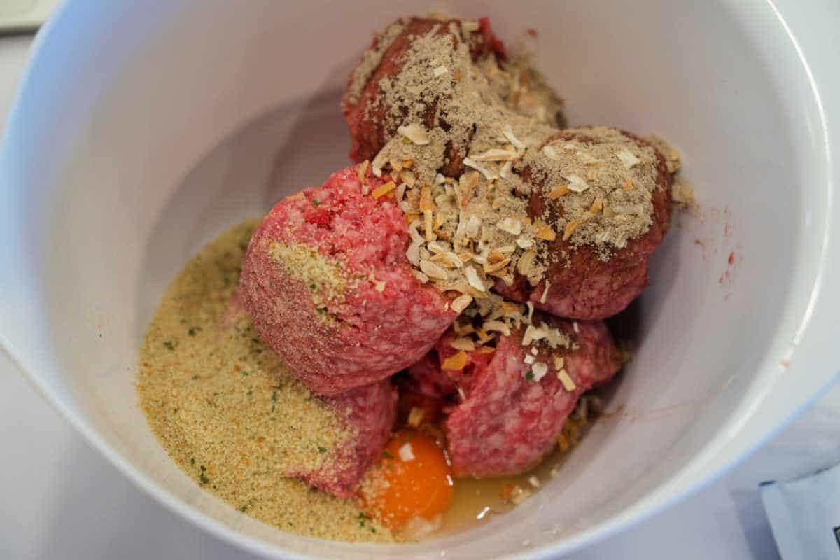 Raw ground meat, breadcrumbs, spices, and a cracked egg in a white mixing bowl, ready to be mixed for delicious meatballs—perfect for pairing with baked spaghetti.