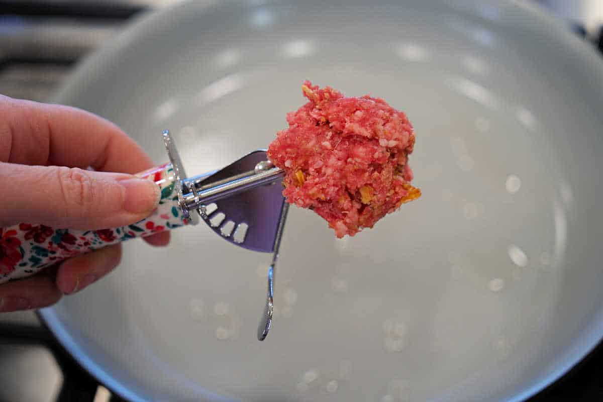 Hand holding a scoop of raw ground meat over an empty frying pan on a stove, perfect for preparing baked spaghetti with meatballs.