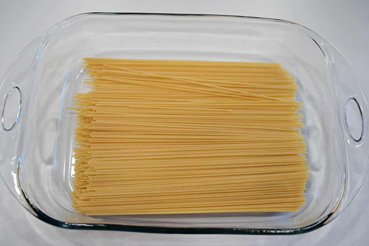 Uncooked spaghetti noodles arranged in a clear glass baking dish on a white surface, ready to be transformed into delicious baked spaghetti with meatballs.