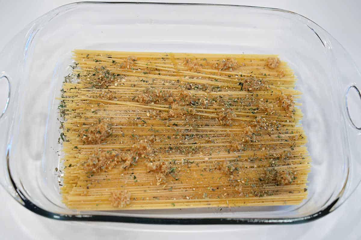 Uncooked spaghetti in a glass baking dish, topped with minced garlic and dried herbs, ready to be transformed into a classic baked spaghetti with meatballs.