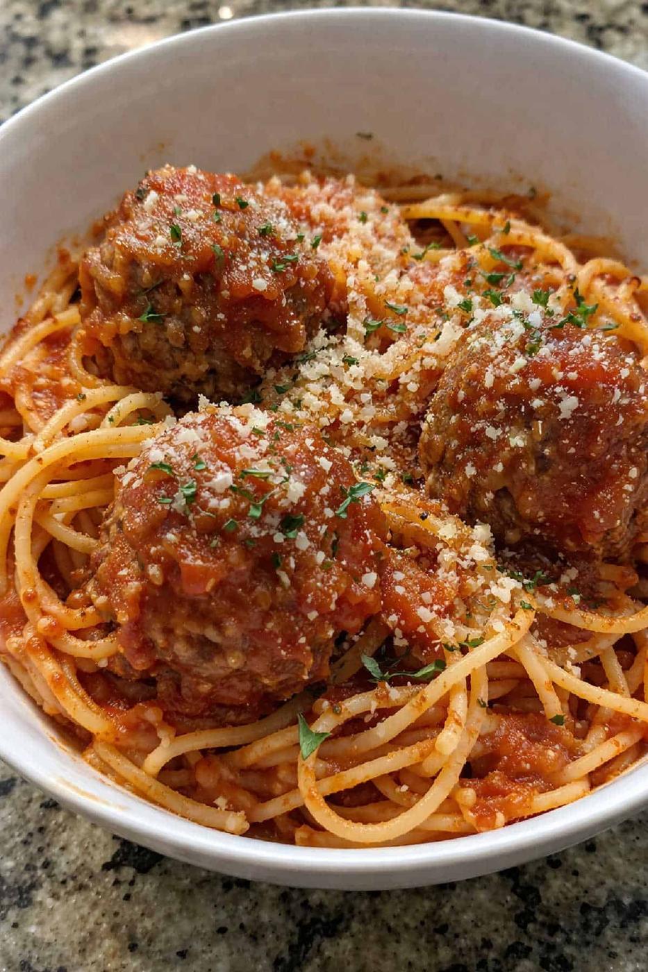 Baked spaghetti with meatballs topped with tomato sauce, grated cheese, and herbs, served in a white bowl on a granite counter.