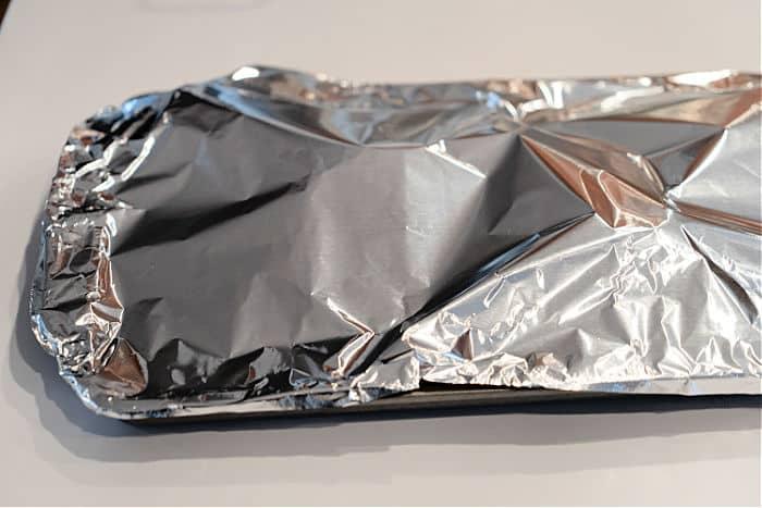 A rectangular baking pan covered tightly with shiny aluminum foil, perfect for preparing delicious baked turkey legs, sits on a white surface.