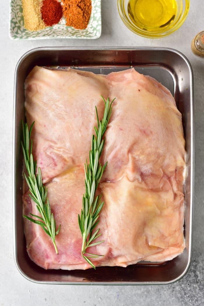 Raw frozen chicken thighs with rosemary sprigs rest in a tray, ready for baking. Nearby, spices and oil await their turn to enhance the flavor. Perfect for transforming frozen chicken thighs into a delicious meal.