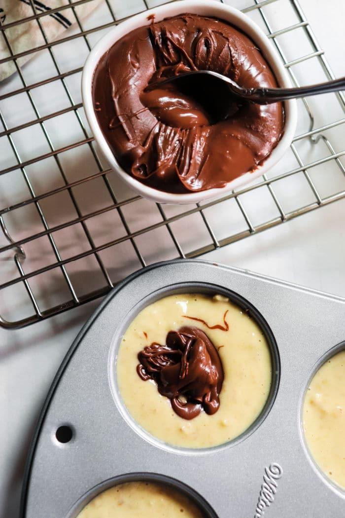 Muffin batter in a pan with a dollop of chocolate, next to a bowl of melted chocolate on a cooling rack, transforms into delightful banana Nutella muffins perfect for any indulgent moment.