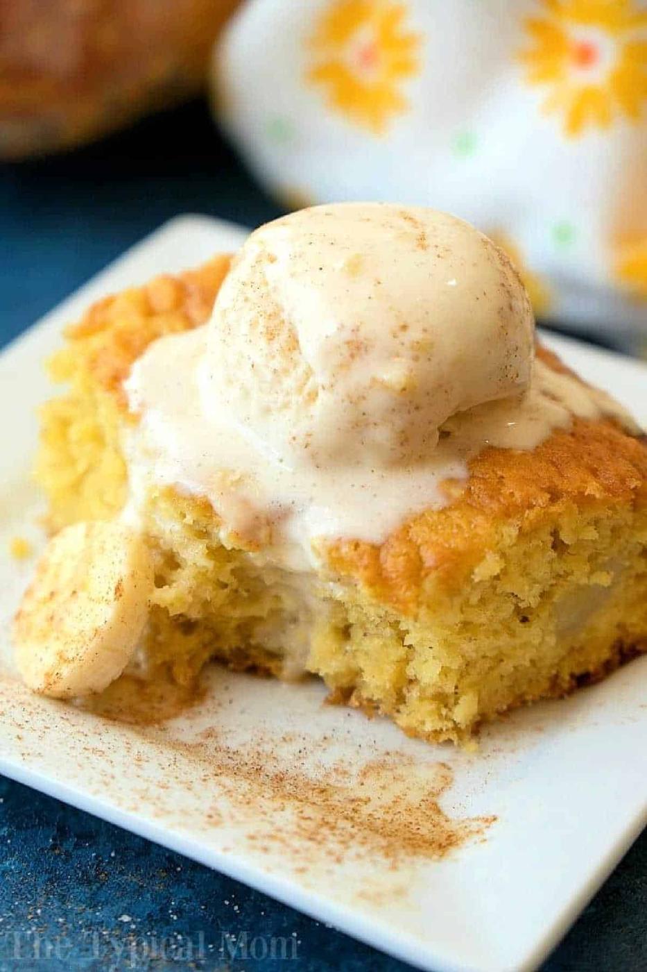 A slice of banana pudding cake topped with melting vanilla ice cream on a white plate, sprinkled with cinnamon, resembles the decadence of a cake mix banana pudding cake.