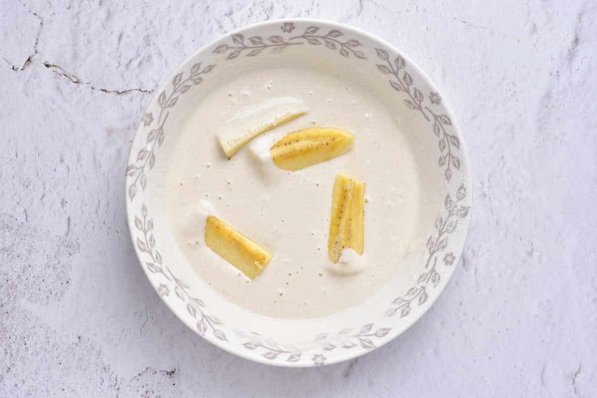 A bowl of tempura is adorned with banana slices, reminiscent of delicate banana tempura, set against a white, textured background.