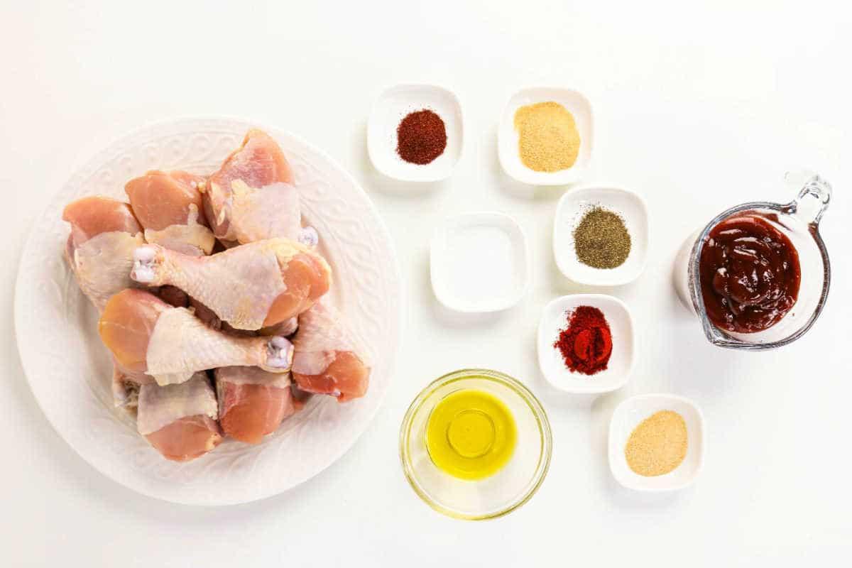 Raw chicken drumsticks on a plate with small bowls of spices, oil, salt, and barbecue sauce on a white background—perfect for your next oven baked BBQ chicken legs recipe.