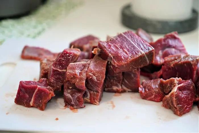 Chopped pieces of raw beef arm roast pieces are placed on a white cutting board, perfect for starting a beef arm roast recipe. The meat is marbled with fat, and the board rests on a kitchen counter. The background features a green-patterned cloth and part of a round, gray object.