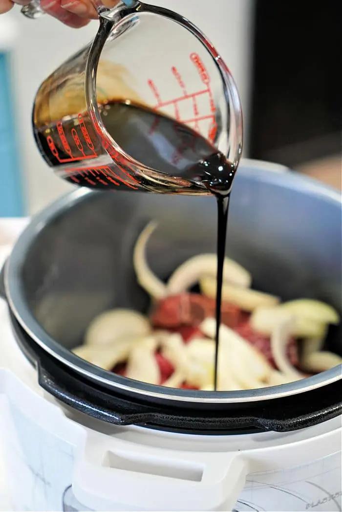 A hand is pouring a balsamic vinegar from a measuring cup into an electric cooker containing sliced onions and beef arm roast. The cooker's lid is open, spotlighting the ingredients as they meld into a mouthwatering recipe.