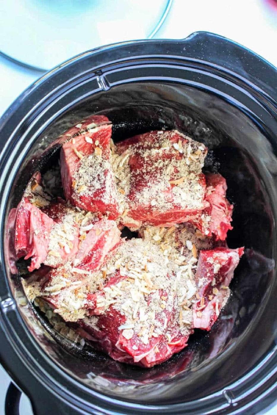 Chunks of raw beef with seasoning in a black slow cooker, ready to be cooked for a hearty Beef Manhattan.