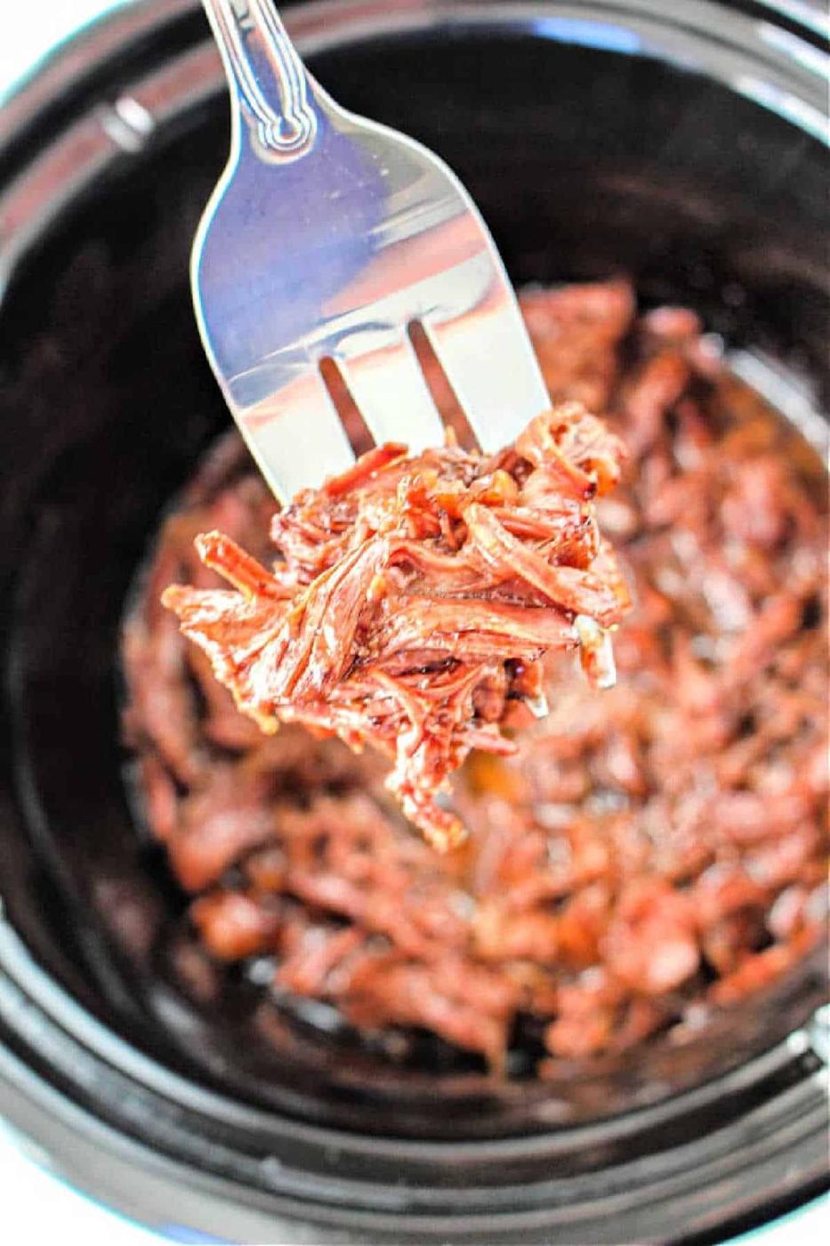 A fork holds shredded Beef Manhattan above a slow cooker filled with more savory, shredded meat.