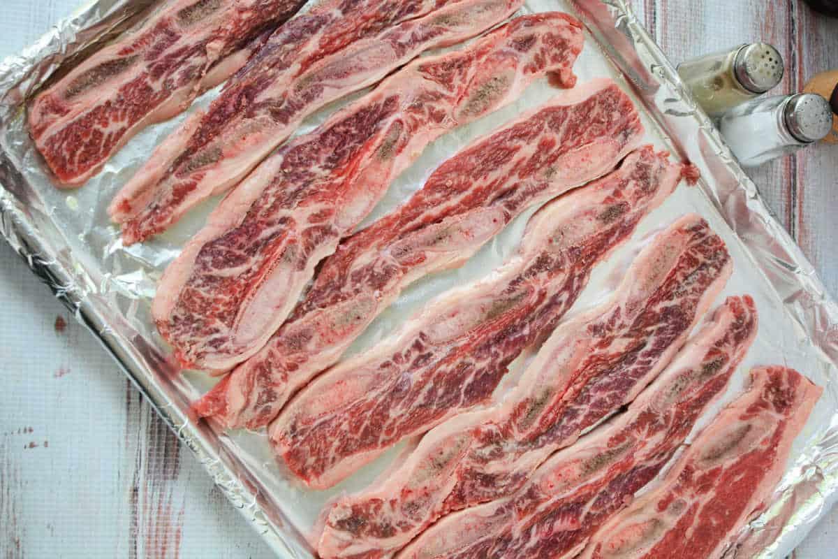 Raw marinated beef riblets arranged on a foil-lined baking sheet, with salt and pepper shakers nearby—perfect for trying out your favorite Beef Riblets Recipe.
