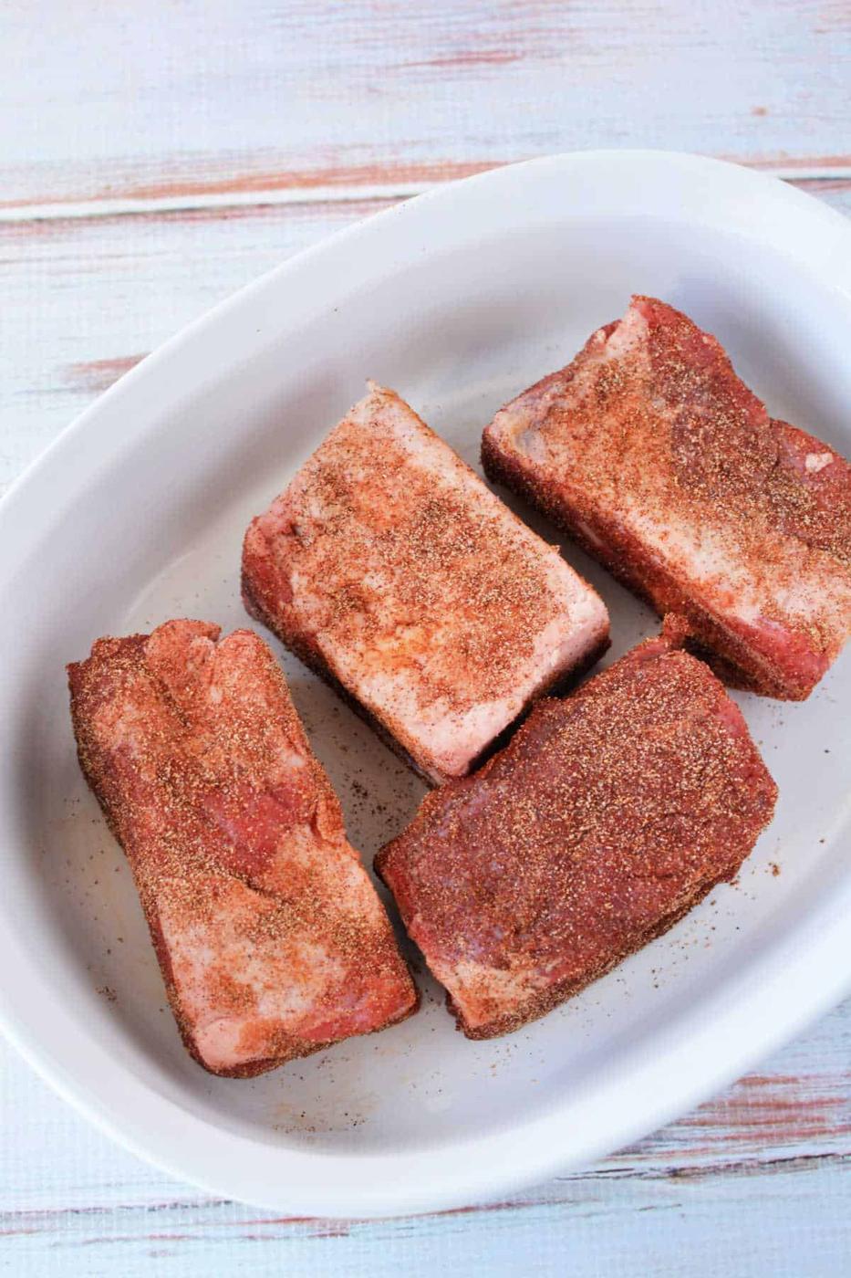 Four seasoned beef ribs in a white dish on a light wooden surface, ready to be compared to the tenderness you achieve with beef ribs in the oven at 275.
