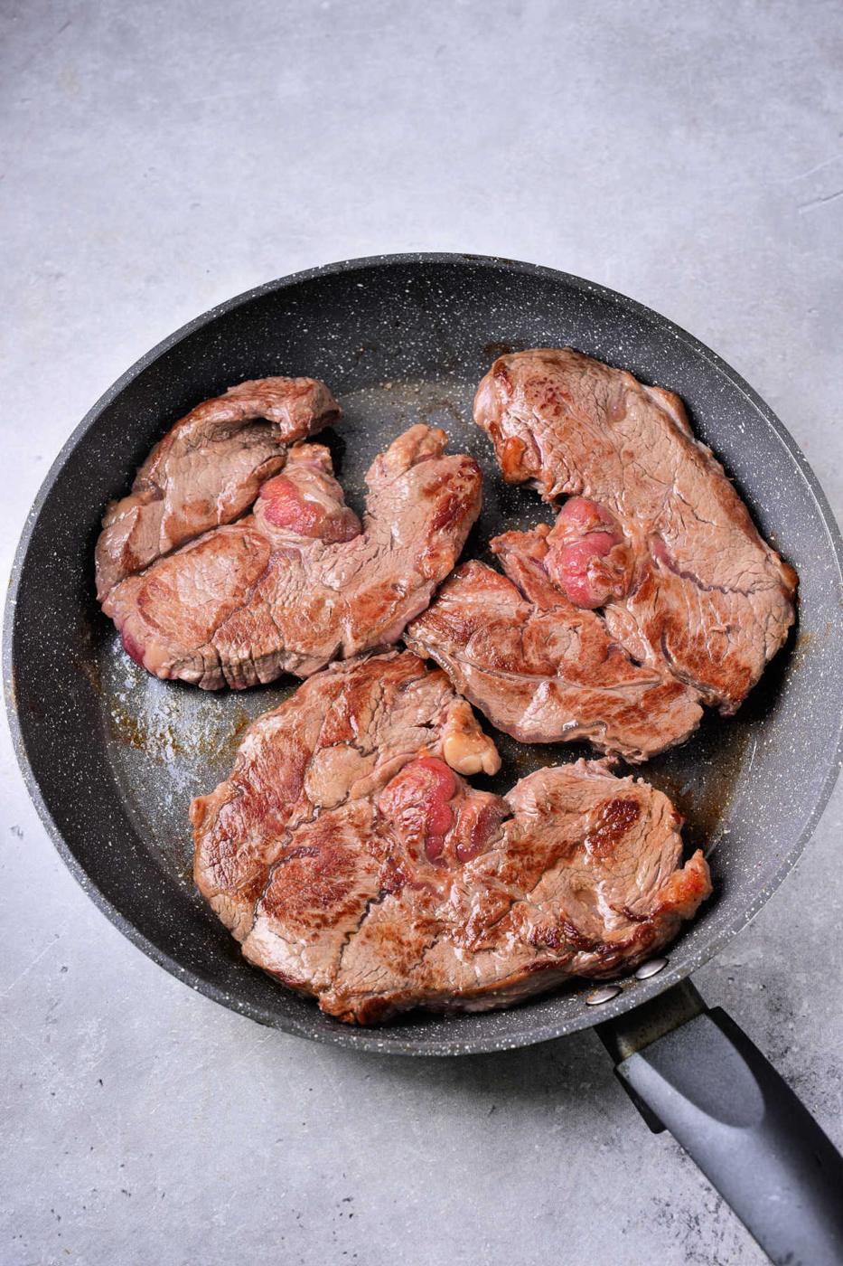 Three pieces of seared steak, perfect for a Beef Shin recipe, cooking in a black frying pan.