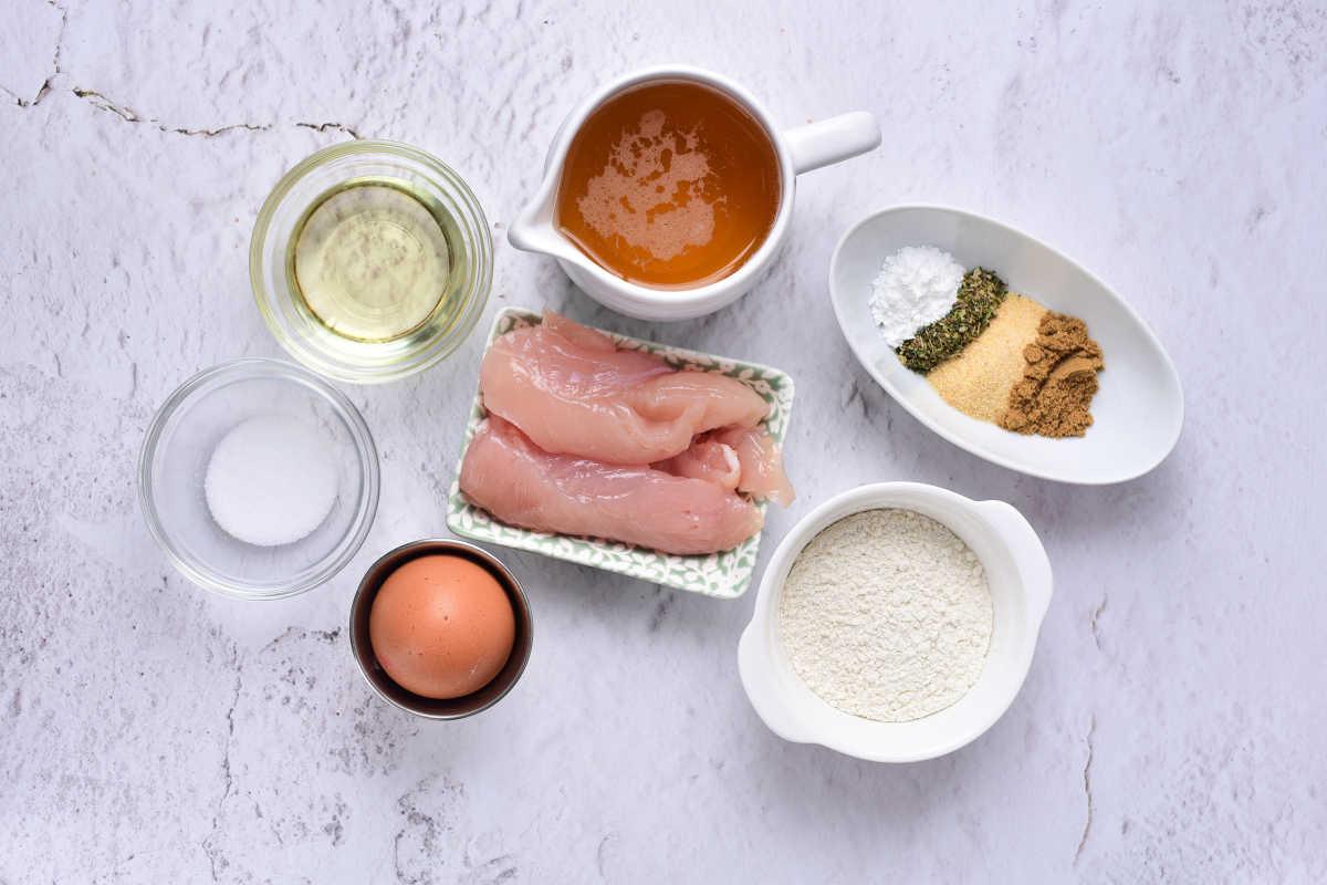 Raw chicken, an egg, flour, seasonings, oil, salt, and broth in bowls on a light textured surface—perfect for making crispy beer battered chicken tenders.