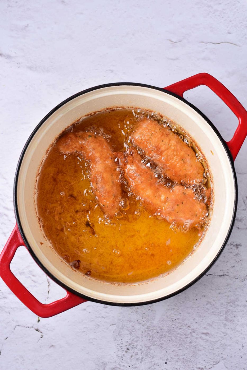 Two beer battered chicken tenders frying in hot oil alongside in a red-handled pot on a light gray surface.