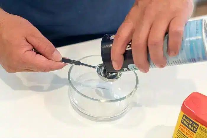 A person pours salt from a container into a measuring spoon over a glass bowl on a white surface, preparing ingredients for the best baked pork chops.