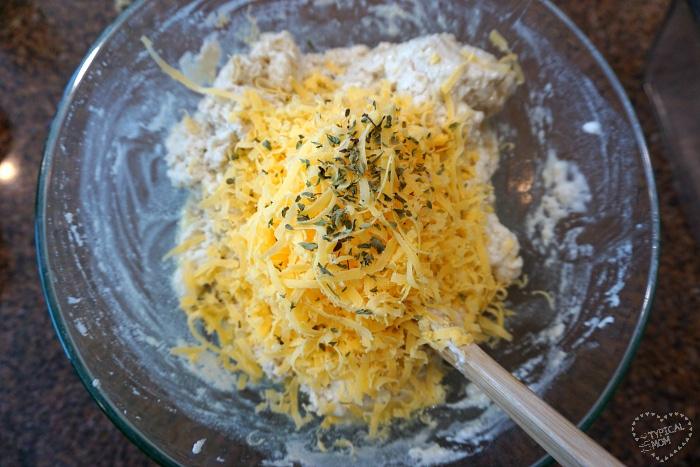 A bowl with shredded cheese and herbs crowns the best beer bread recipe. A wooden spoon rests elegantly in the dough mixture, ready to craft this delicious masterpiece.