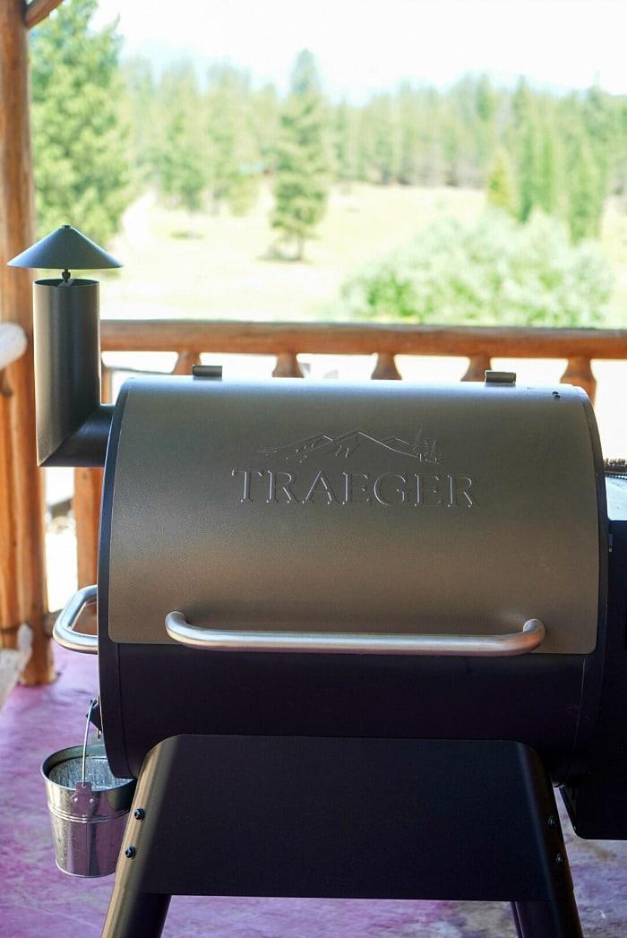 A Traeger grill is positioned on a wooden porch with a serene forest background.