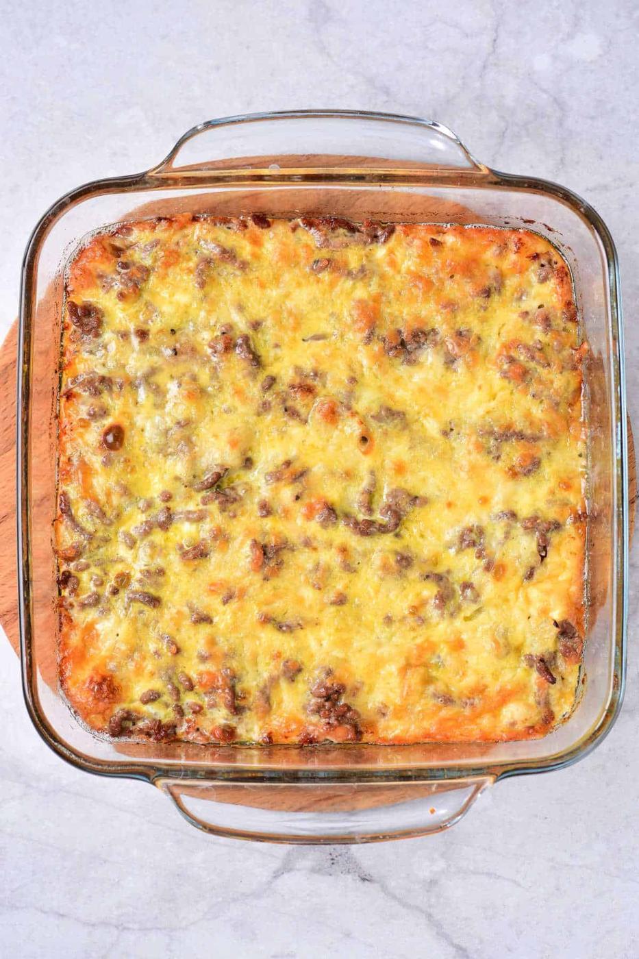 A Big Mac Casserole with a golden-brown cheese topping sits in a clear glass baking dish. The casserole, featuring ground meat and layers of melty cheese, boasts a slightly crispy surface. It rests invitingly on a white marble countertop, promising classic flavors in every bite.