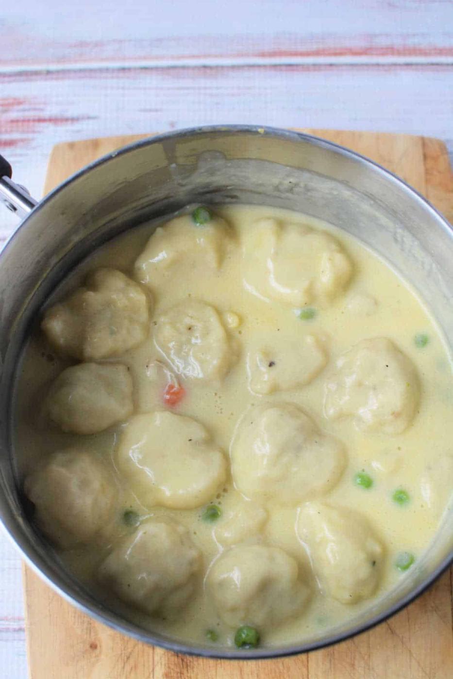 A pot of creamy chicken and dumplings made with Bisquick, peas, and carrots sits on a wooden cutting board.