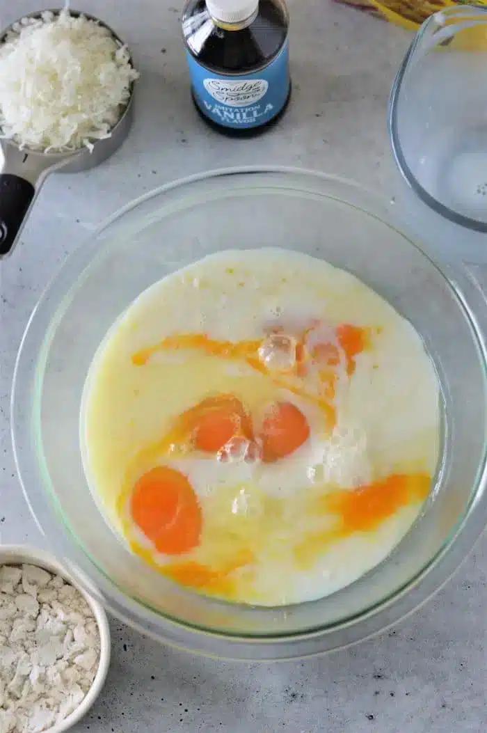 A mixing bowl contains eggs, milk, and other liquid ingredients for a Bisquick Impossible Pie Recipe. Nearby are a measuring cup with white shredded coconut, a bottle of vanilla extract, and a small bowl of flour on a gray countertop.