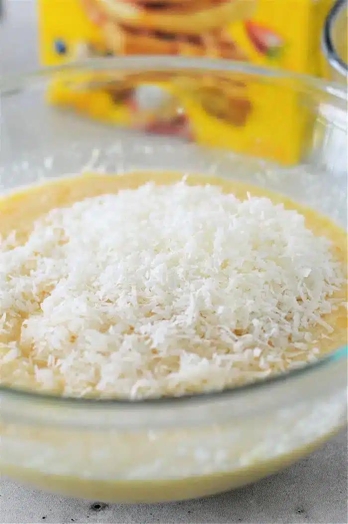 A glass bowl filled with Bisquick Impossible Pie batter mixture, topped with shredded coconut. A box in the background is blurry and suggests ingredients for baking.