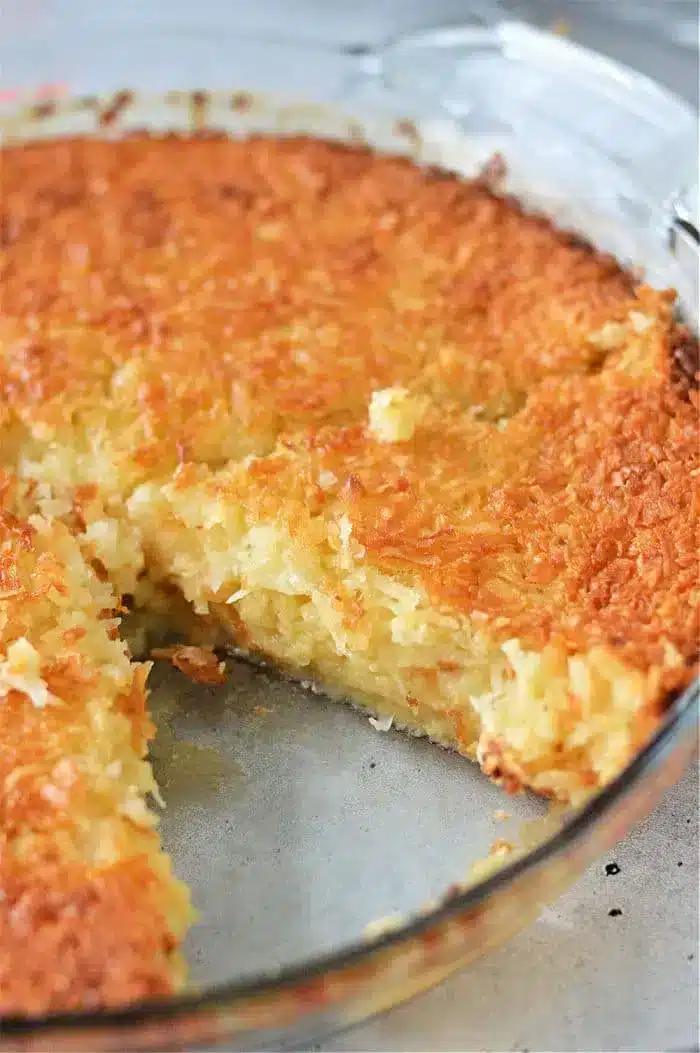 A close-up of a coconut pie, reminiscent of a Bisquick Impossible Pie Recipe, in a round glass dish with a slice removed, revealing its creamy interior. The top is golden brown and textured with shredded coconut. The pie rests on a grey surface.