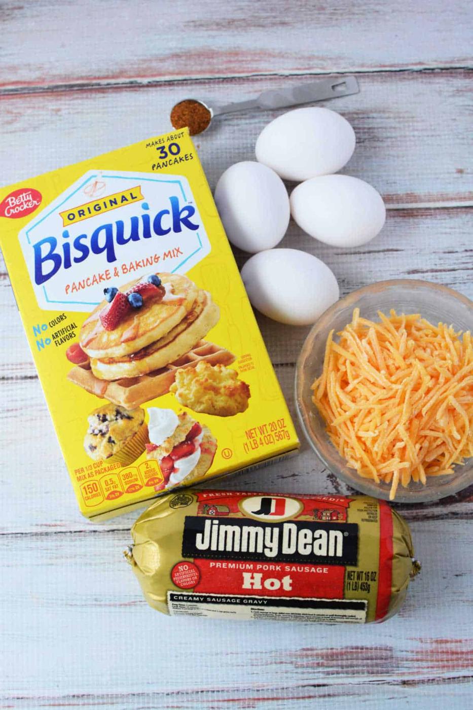 A Bisquick box, three eggs, shredded cheese, a hot sausage package, and a teaspoon of spice sit ready on a white wooden surface, perfect for whipping up savory muffins.