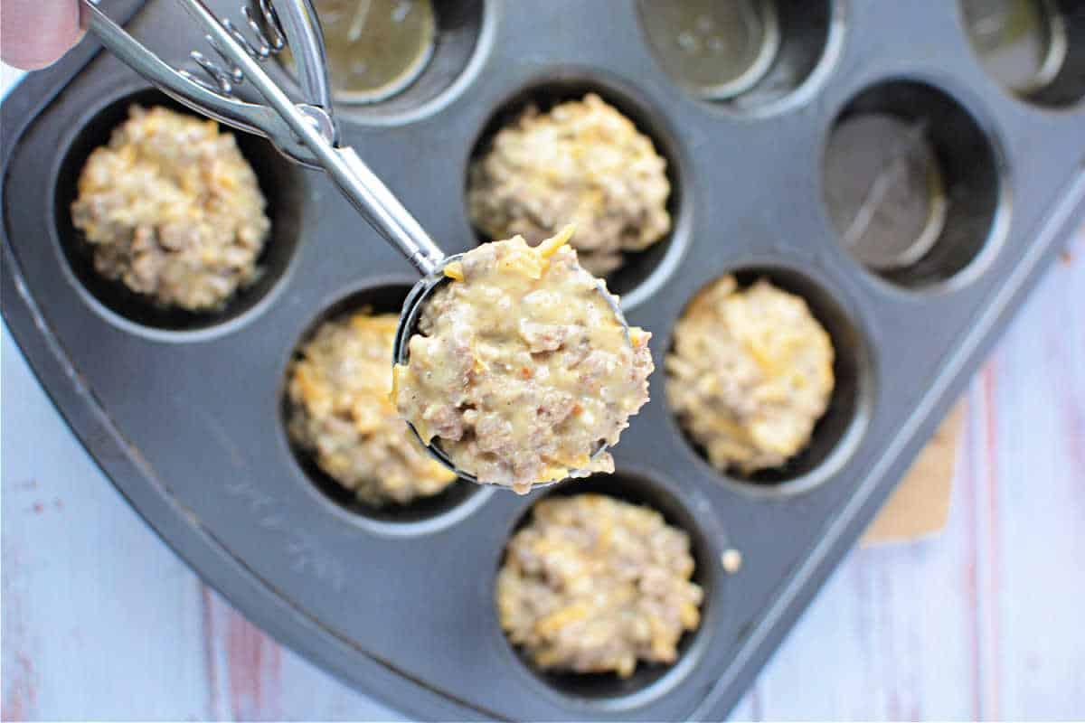 A scoop of muffin batter overflows a muffin pan filled with delicious muffins, ready to bake on a light wooden surface, much like preparing a savory Bisquick sausage treat.