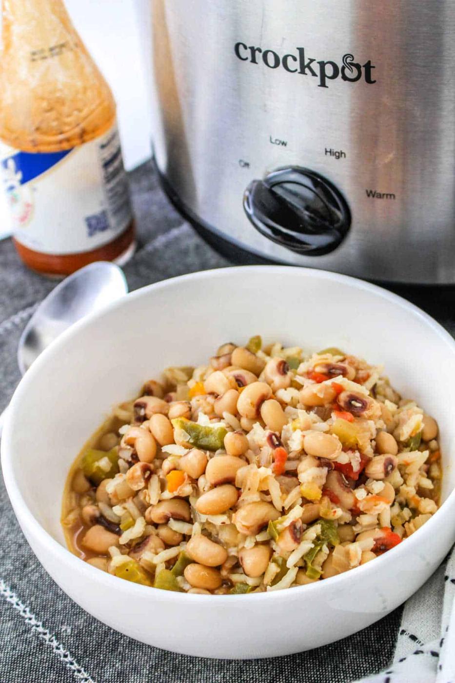 A bowl of Black Eyed Peas and Rice sits in front of a crockpot, accompanied by a spoon and a bottle of sauce, ready to enjoy.
