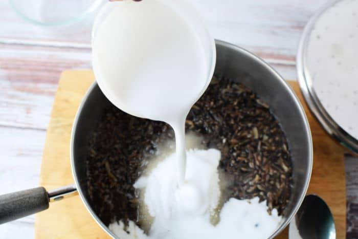 Pouring creamy coconut milk into a pot filled with black rice and sugar on a wooden surface, creating a luscious black rice pudding.
