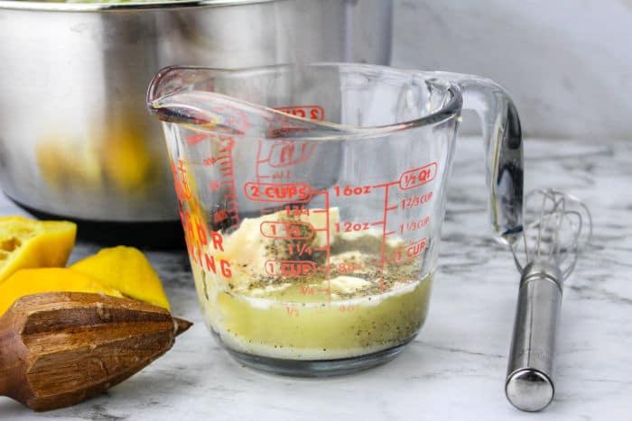 A glass measuring cup with coleslaw dressing sits on a marble surface, accompanied by a whisk, lemons, and a citrus reamer—perfect for preparing tangy mixtures like Blue Cheese Coleslaw.