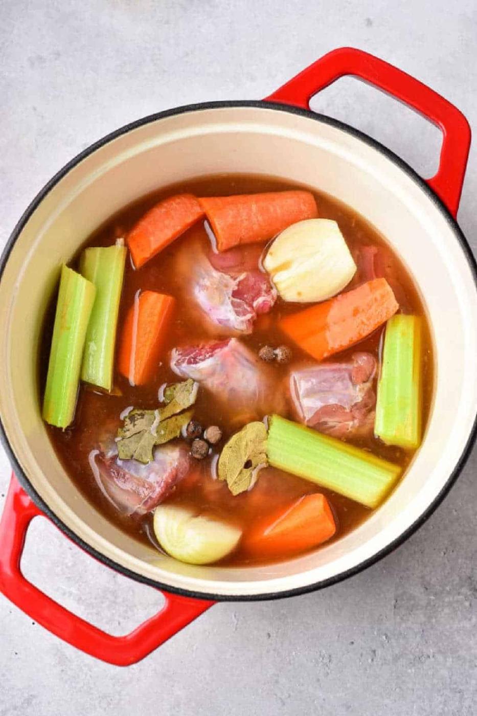 Red pot with raw beef, carrots, celery, onions, bay leaves, and broth—perfect for making soups or pairing with boiled turkey necks—ready to cook on a gray surface.