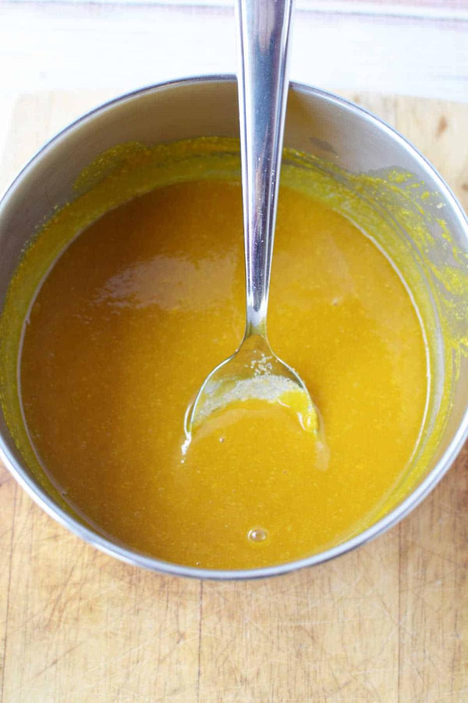 A metal bowl of yellow-orange Bologna Burnt Ends sauce with a spoon resting inside sits on a wooden surface.