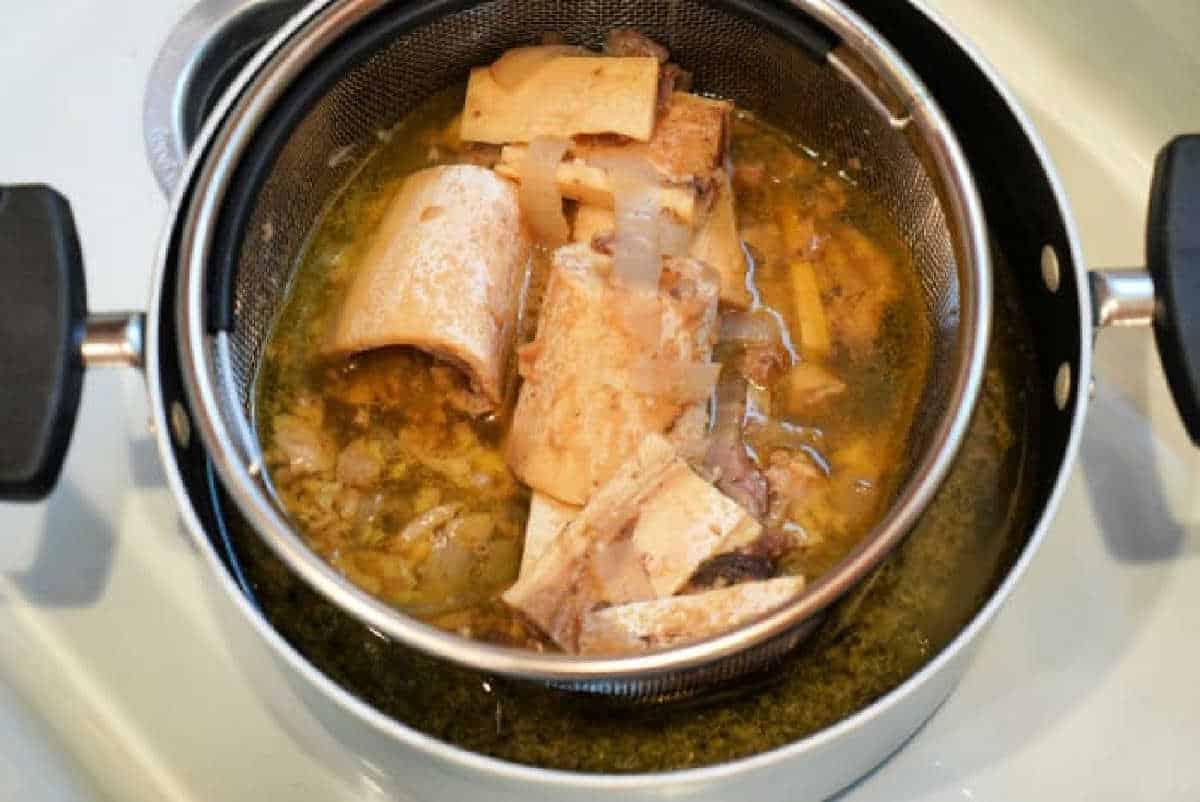 Large beef bones and vegetables in a strainer over a pot of broth on the stovetop—perfect for preparing a nourishing Bone Broth Recipe Instant Pot enthusiasts will love.