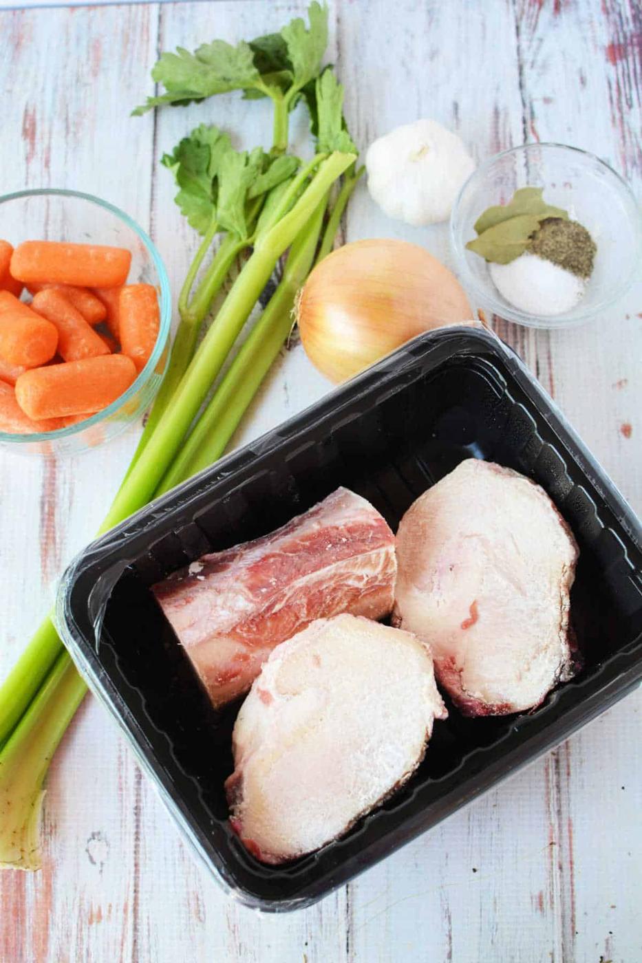 Ingredients for bone broth: celery, carrots, garlic, and onion lay next to beef bones on a tray. A bowl of spices with bay leaves completes the setup. Perfectly suited for a slow cooker, these elements promise depth and warmth in every sip.