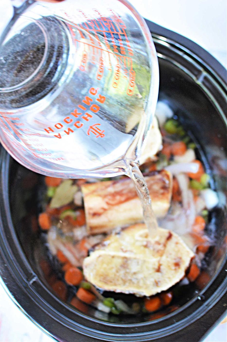 Pouring water into a slow cooker, creating a rich bone broth with vegetables and bones.