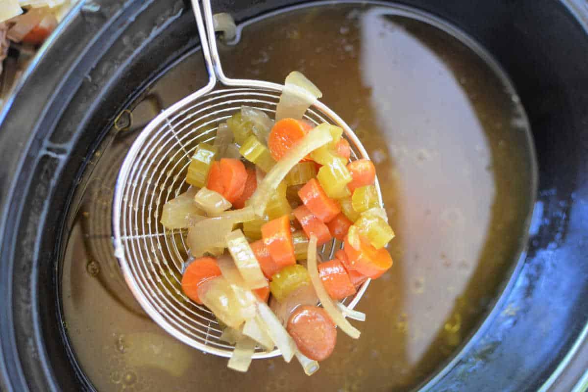 A wire ladle lifts diced carrots, celery, and onions above a pot of simmering bone broth, creating a fragrant medley perfect for your slow cooker.