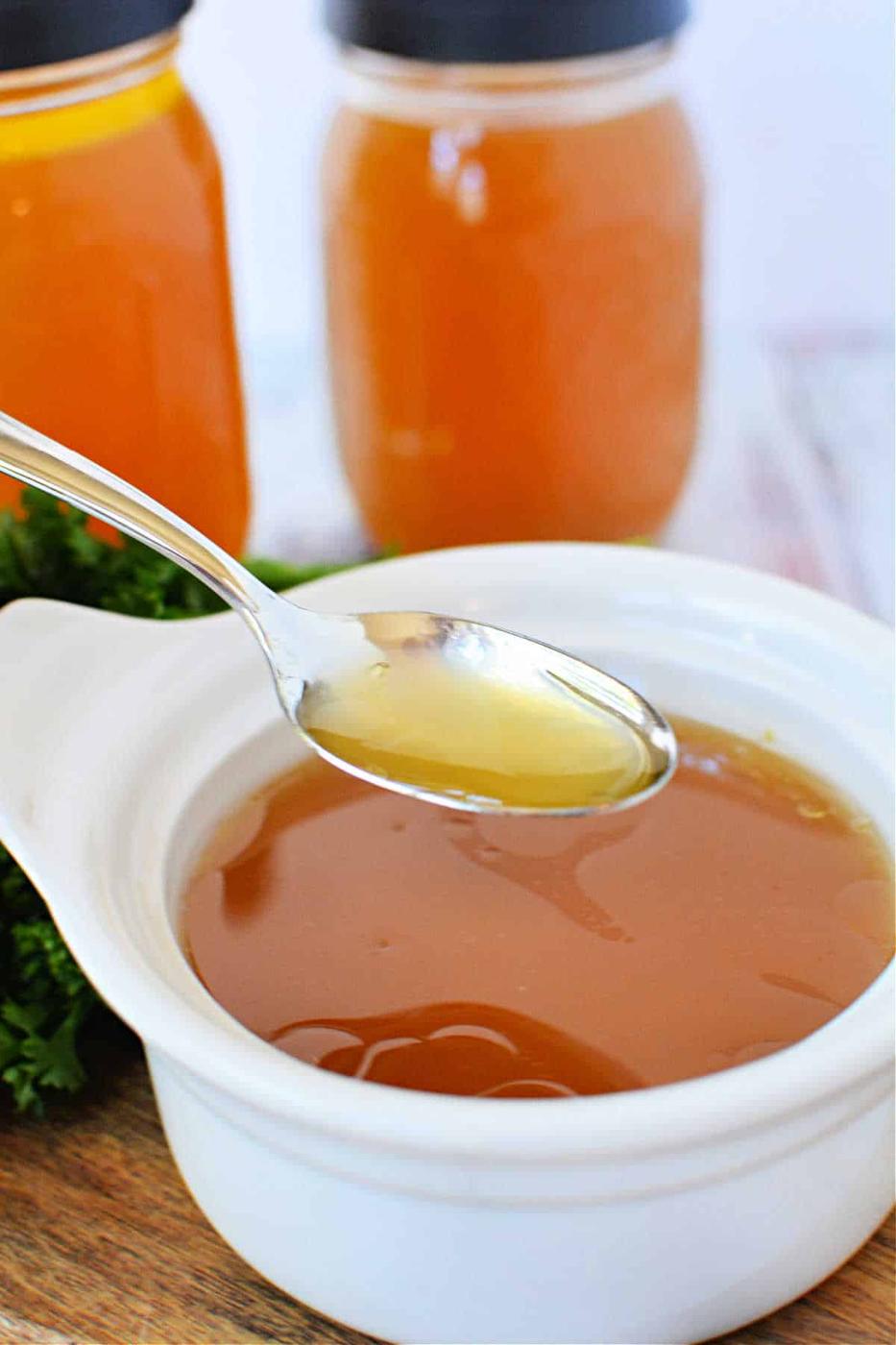 A spoon hovers over a white bowl, filled with homemade bone broth made in a slow cooker, with two jars in the background.